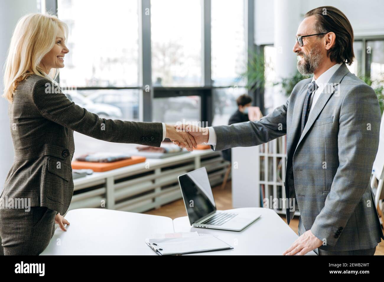 Successful middle aged colleagues are standing in modern office, coming to agreement at conference. Satisfied business partners shaking hands after negotiations Stock Photo