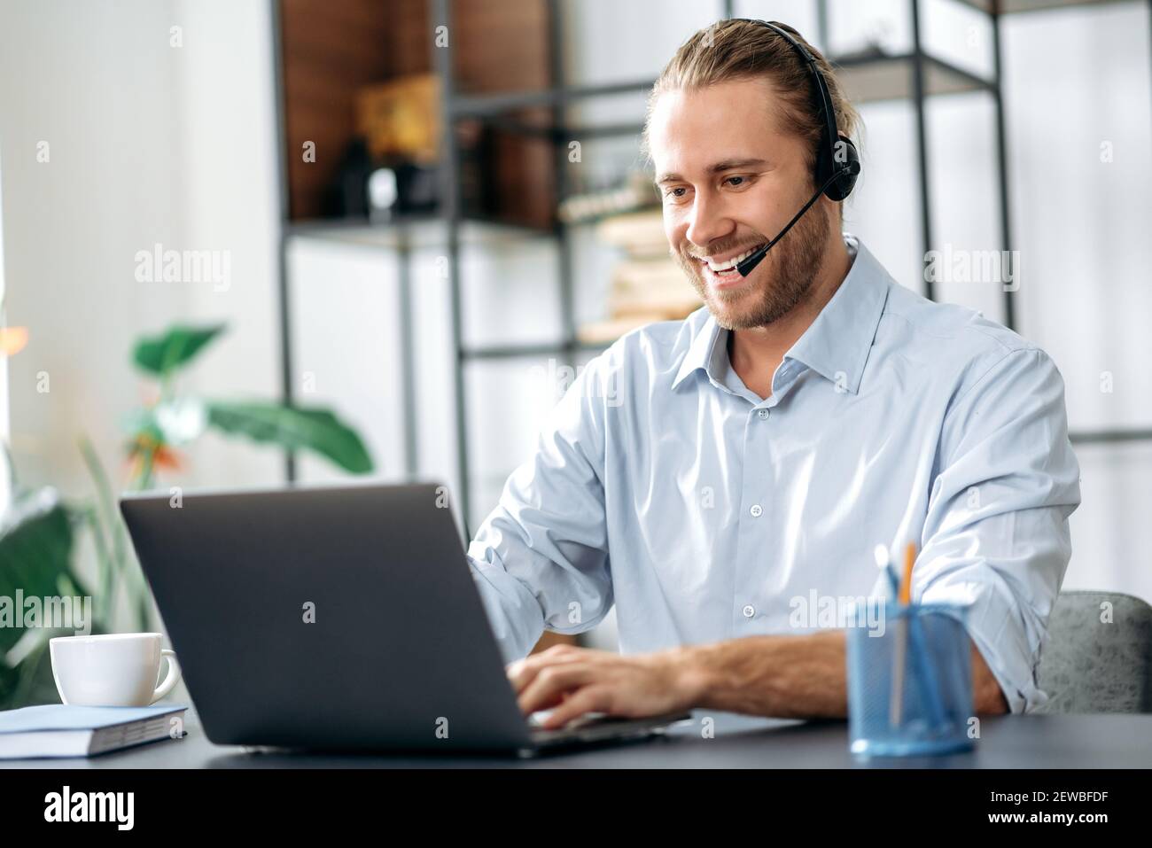 Joyful young adult male manager or call center worker in headset is looks at the laptop screen, having online business meeting. Successful employee talking online with colleagues Stock Photo