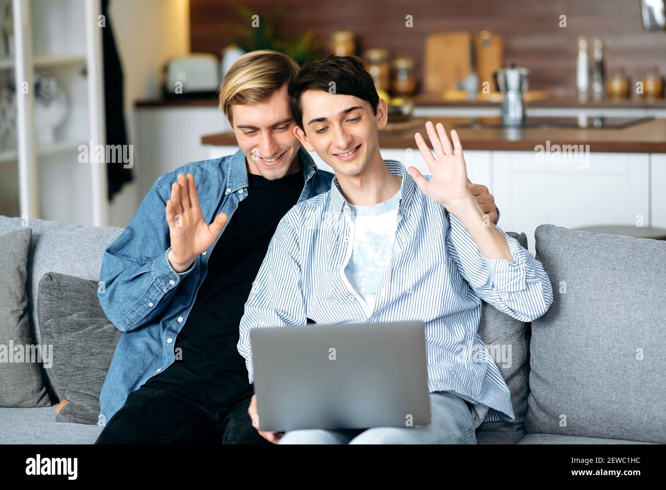 Homosexual couple. Online communication. Gay couple relax on the couch in the living room, use a laptop, they chatting with friends by video call, smiling, waving hands, greeting Stock Photo