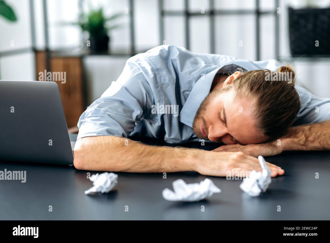Tired caucasian freelancer guy or student is fall asleep at the work desk. Exhausted busy man working overtime, sits at the desk at home, need a rest, overwork concept. Work from home Stock Photo