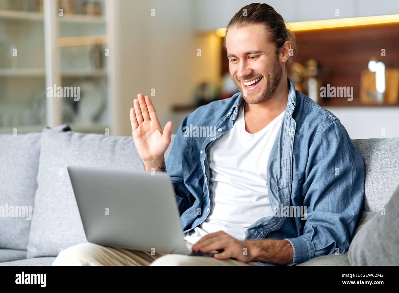 Joyful male freelancer using laptop, have a video call, communicate with colleagues or friends by video conference, waving hand and smiling. Handsome caucasian man is sitting on a sofa, working from Stock Photo