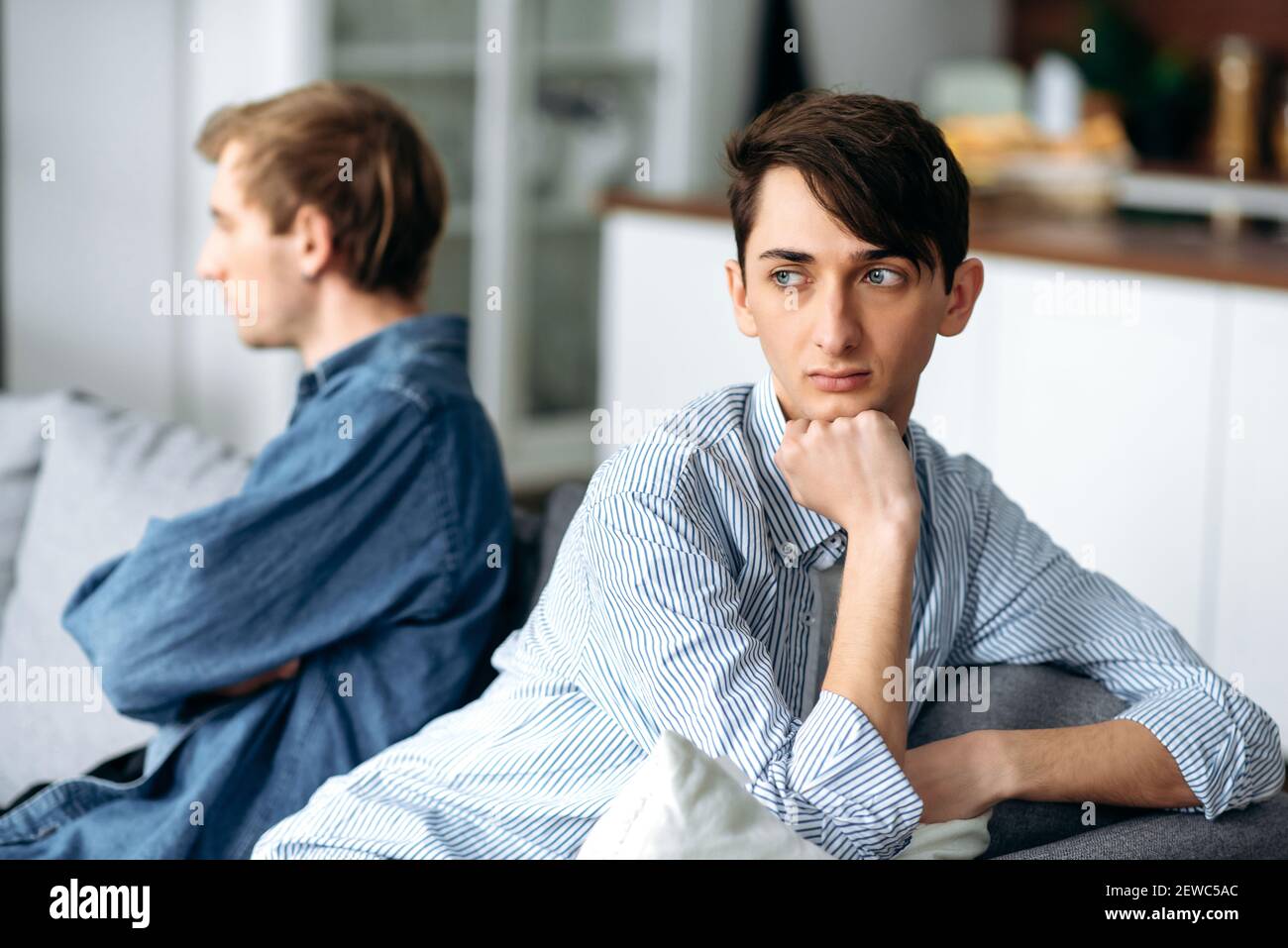 Quarrel between a gay couple in love. An upset guy and his partner are offended and ignore each other while sitting on the couch, turning away in different directions Stock Photo