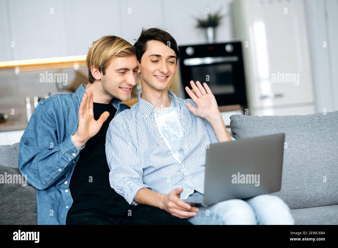 Homosexual couple. Online communication. Gay couple relax on the couch in the living room, use a laptop, they chatting with friends by video call, smiling, waving hands, greeting Stock Photo