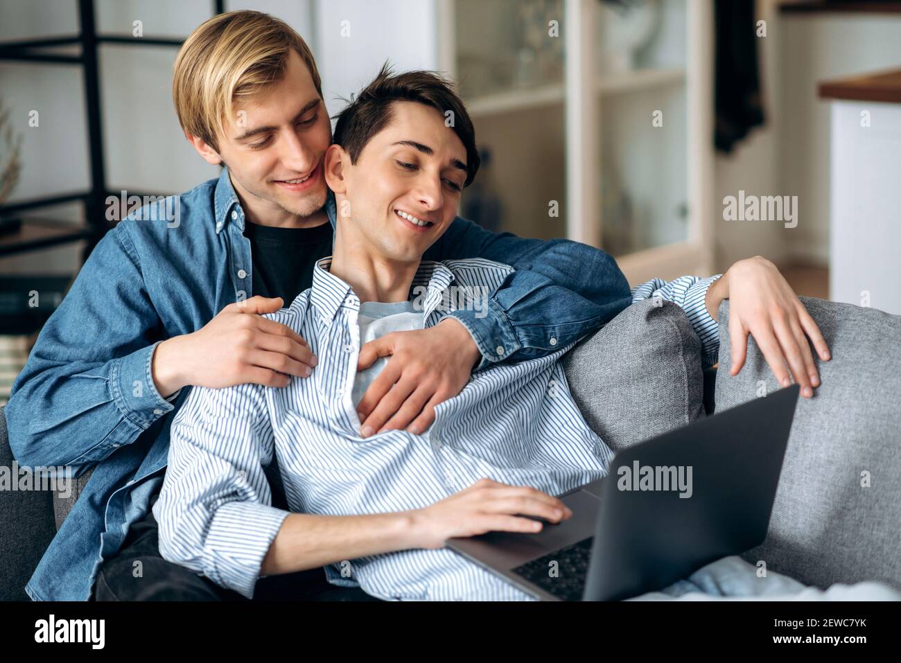Homosexual couple. Gay men relax on the couch in the living room, use a laptop, browse the Internet, watch a movie, shopping online, spend time together Stock Photo