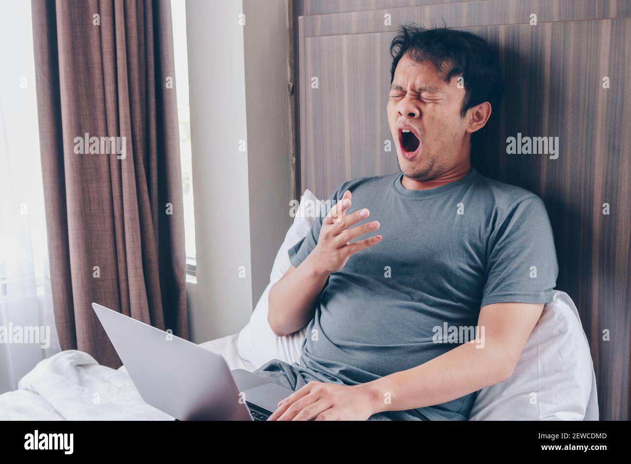Tired stressed young Asian man feeling sleepy and tired while using laptop on the bed in bedroom. Hard work concept. Stock Photo