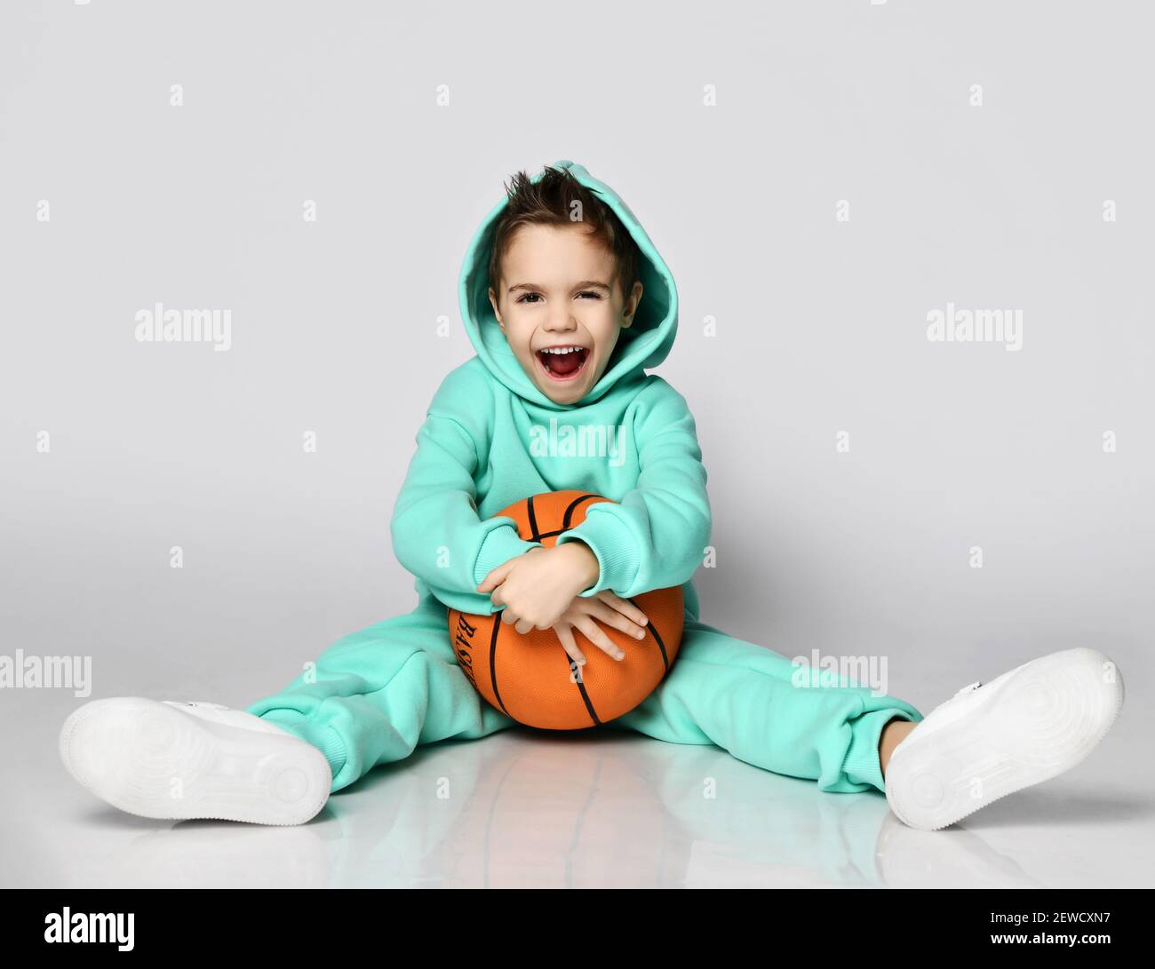 Active frolic boy in modern green, mint color sportswear hoodie and pants sits on floor with basketball and his hood on Stock Photo