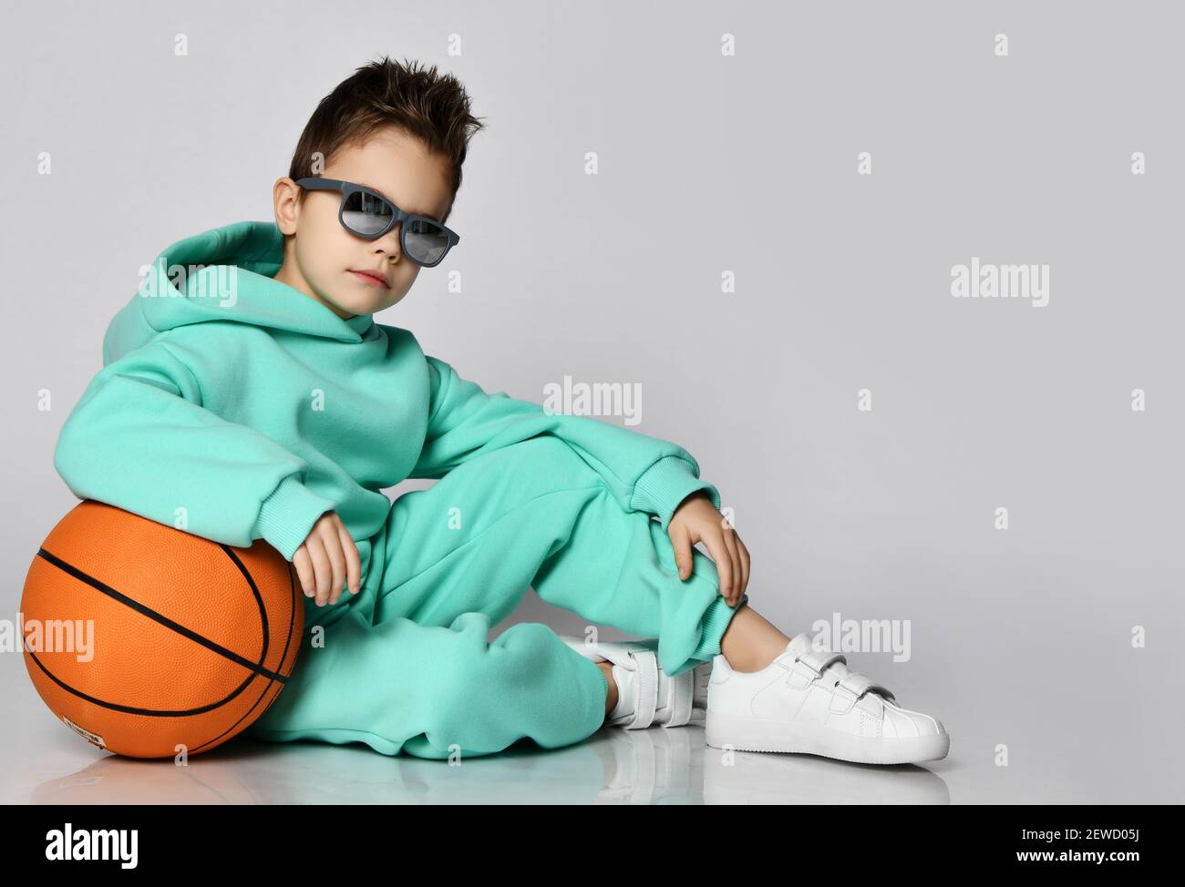 Cool guy schoolboy in modern green, mint color sportswear hoodie, pants and white sneakers sits on floor with basketball Stock Photo