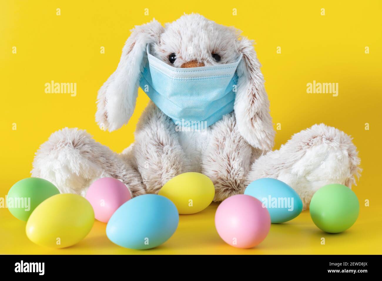 Easter stuffed toy bunny in protective face mask and colored eggs on yellow background. Ester holiday during coronavirus pandemic concept. Stock Photo