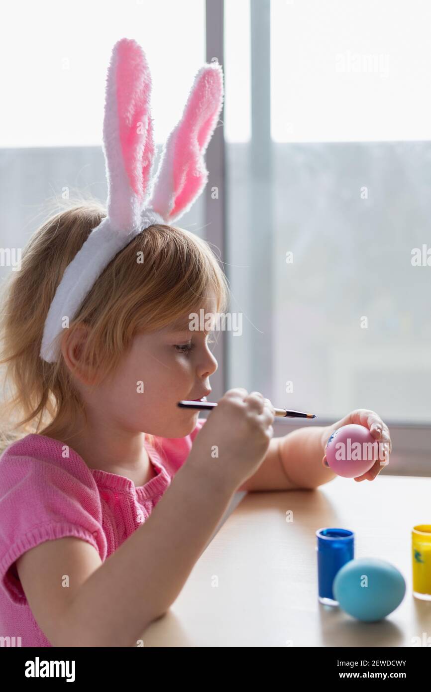 Little girl in Easter bunny ears painting colored eggs. Easter family holiday celebration at home and craft concept. Stock Photo