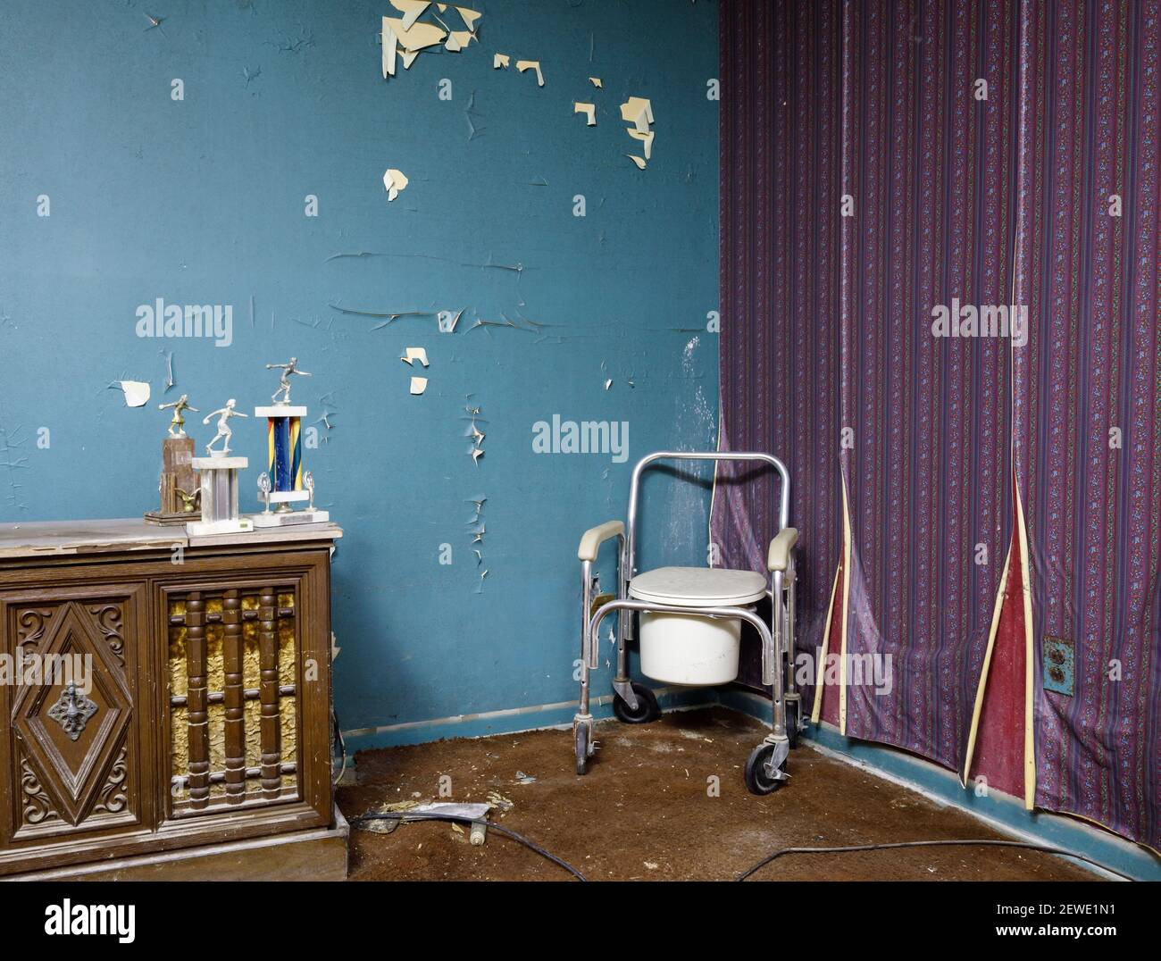 A bedside commode on wheels in an abandoned room with bowling trophies on top of a hutch inside an abandoned home. Ontario, Canada. Stock Photo
