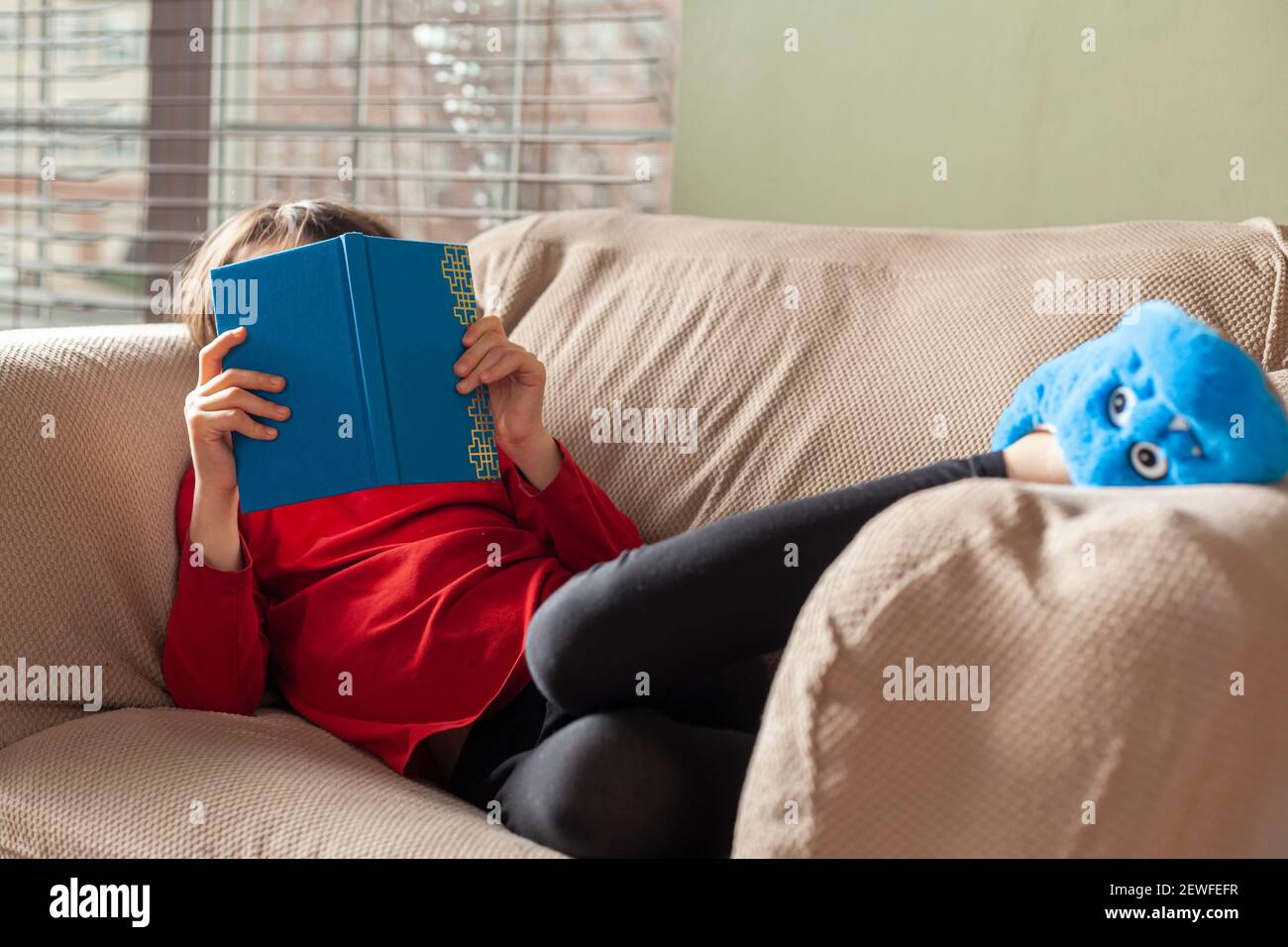 A cute little caucasian girl wearing casual blue slipper socks, leggings and a red sweater is lying on an armchair and reading a book in front of the Stock Photo