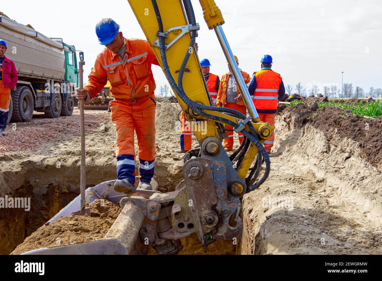 Worker is leap out from quadratic trench with little help of digger's bucket full of excavated ground. Stock Photo