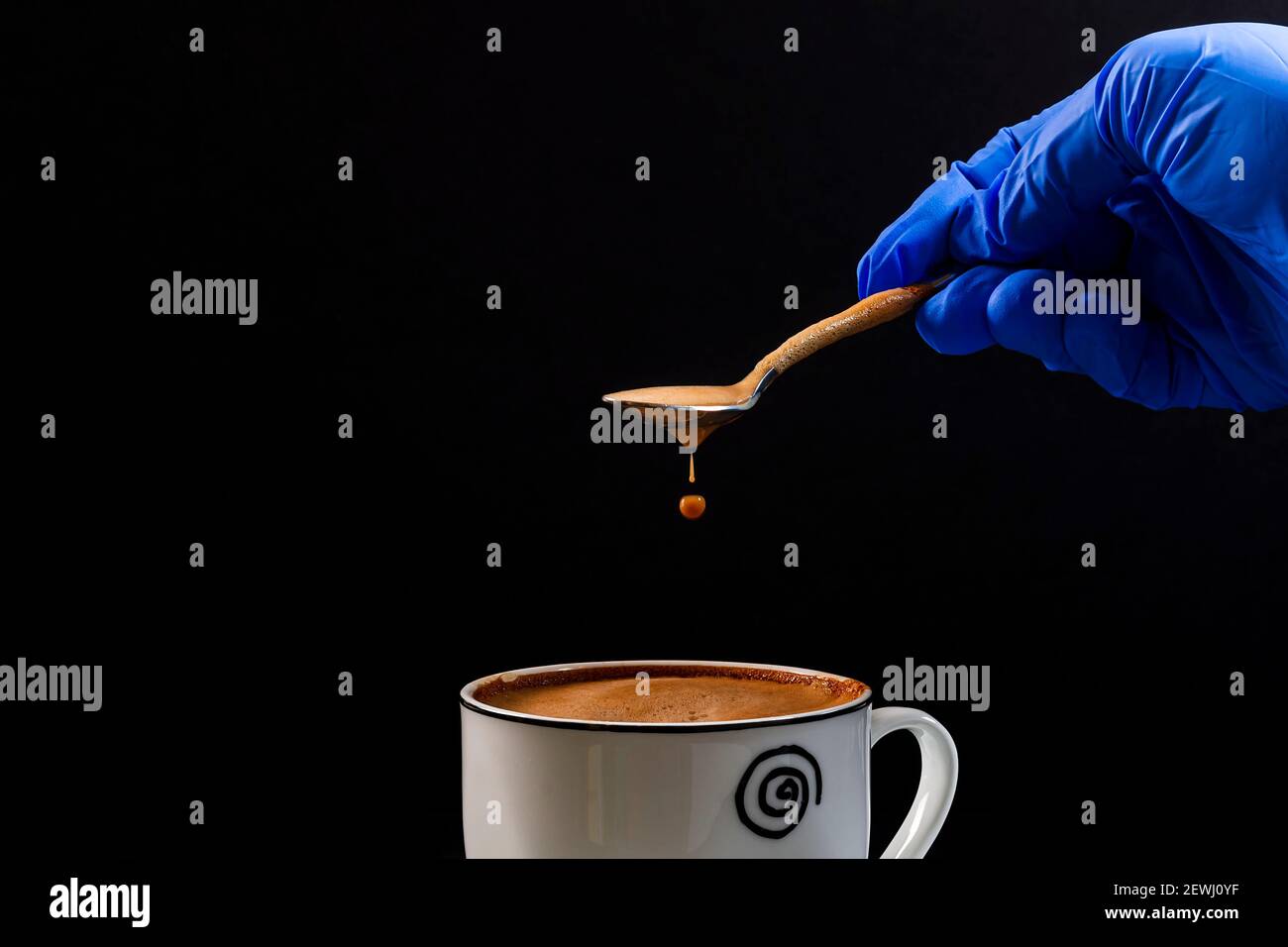 The hand of a person wearing a latex glove holds a spoon from which a drop of latte falls.This is a horizontal format photo taken against a black back Stock Photo