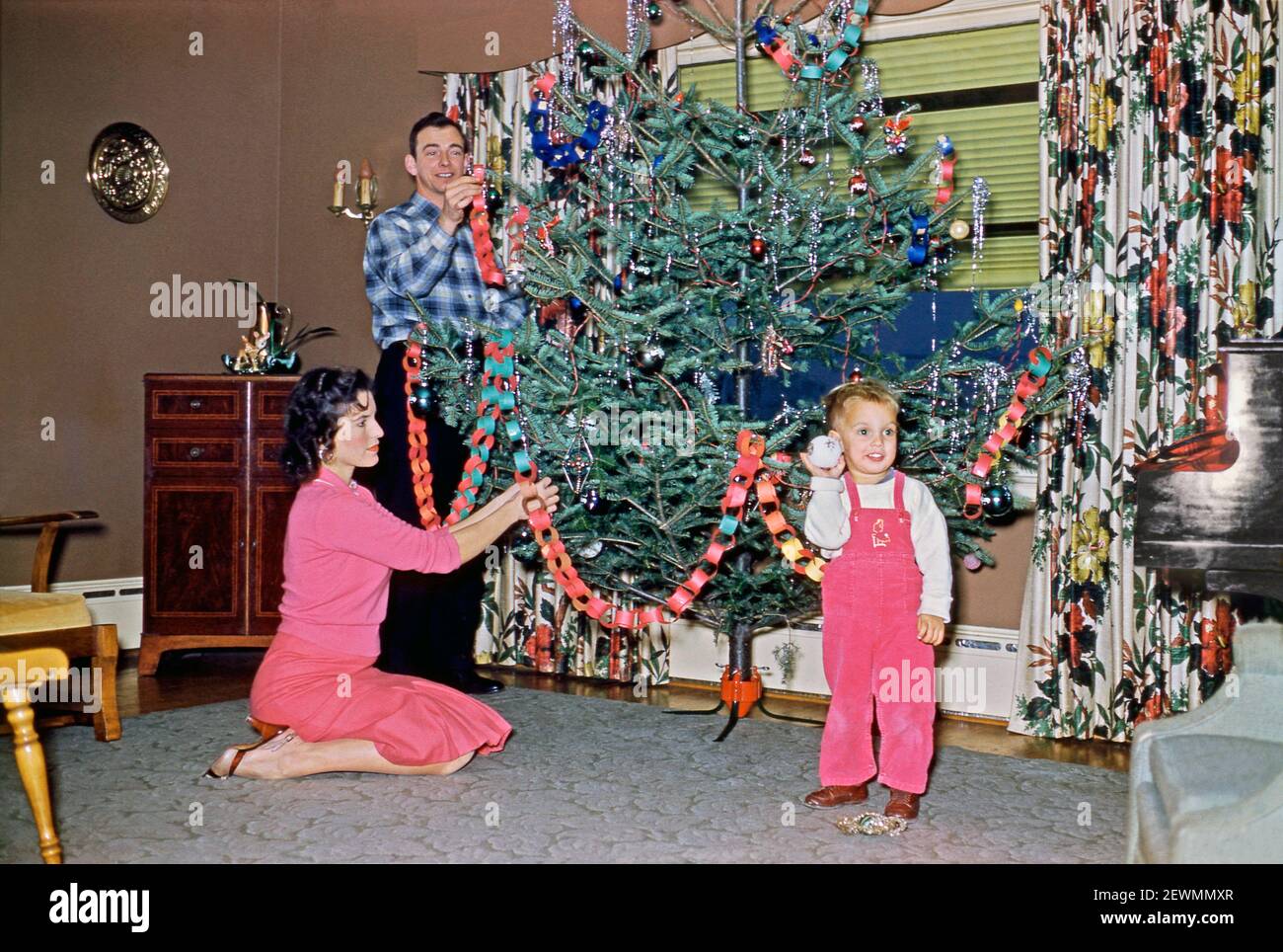 Putting Christmas decorations on the tree, in a living room in the USA in 1958. The traditional decorations for the large tree include tinsel, linked paper garlands and baubles, one of which the young boy is holding. This image is from an old American amateur Kodak colour transparency – a vintage 1950s photograph. Stock Photo