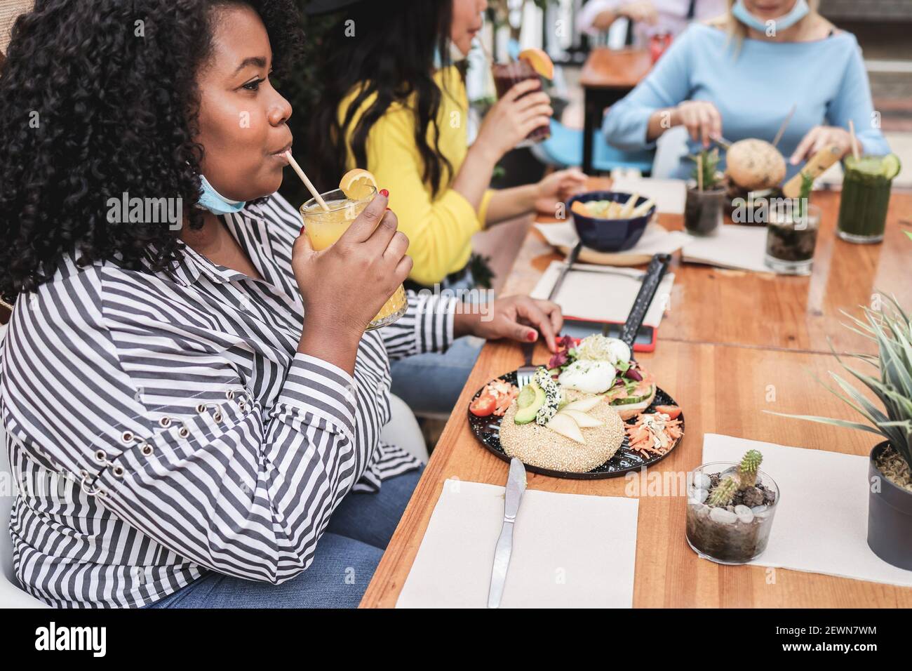 Young multiracial friends having breakfast in restaurant outdoors with masks under chins - Focus on african girl Stock Photo