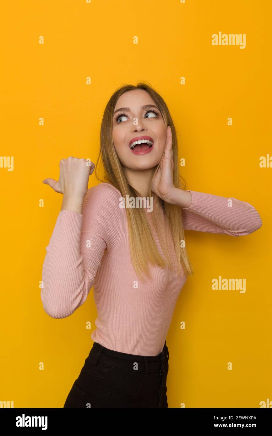 Excited young woman in pink sweater is pointing behind herself with thumb, looking away and talking. Side view. Waist up studio shot on yellow backgro Stock Photo