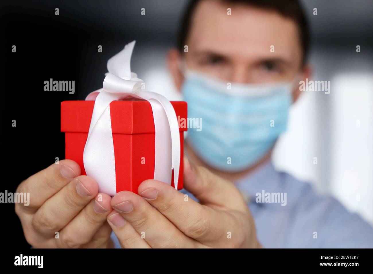 Red gift box in male hand close up, celebration during coronavirus pandemic concept. Man medical face mask and office clothes, selective focus on gift Stock Photo