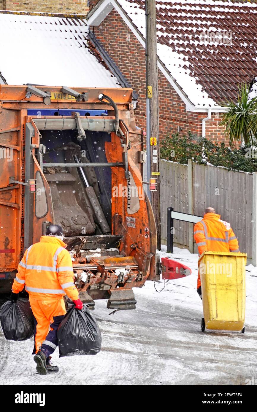Ice snow & cold winter weather high visibility refuse collector workers handling yellow wheelie bin & rubbish sacks at back of dustcart on icy road UK Stock Photo