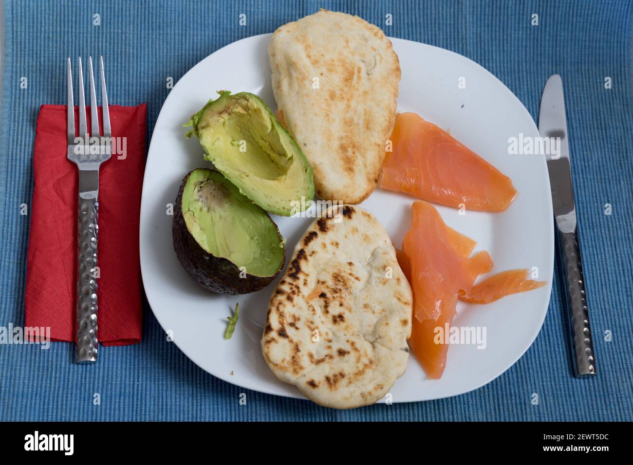 two naan breads toasted with salmon and half avocado on a white plate , red napkin, fork and knife , on blue table mat Stock Photo