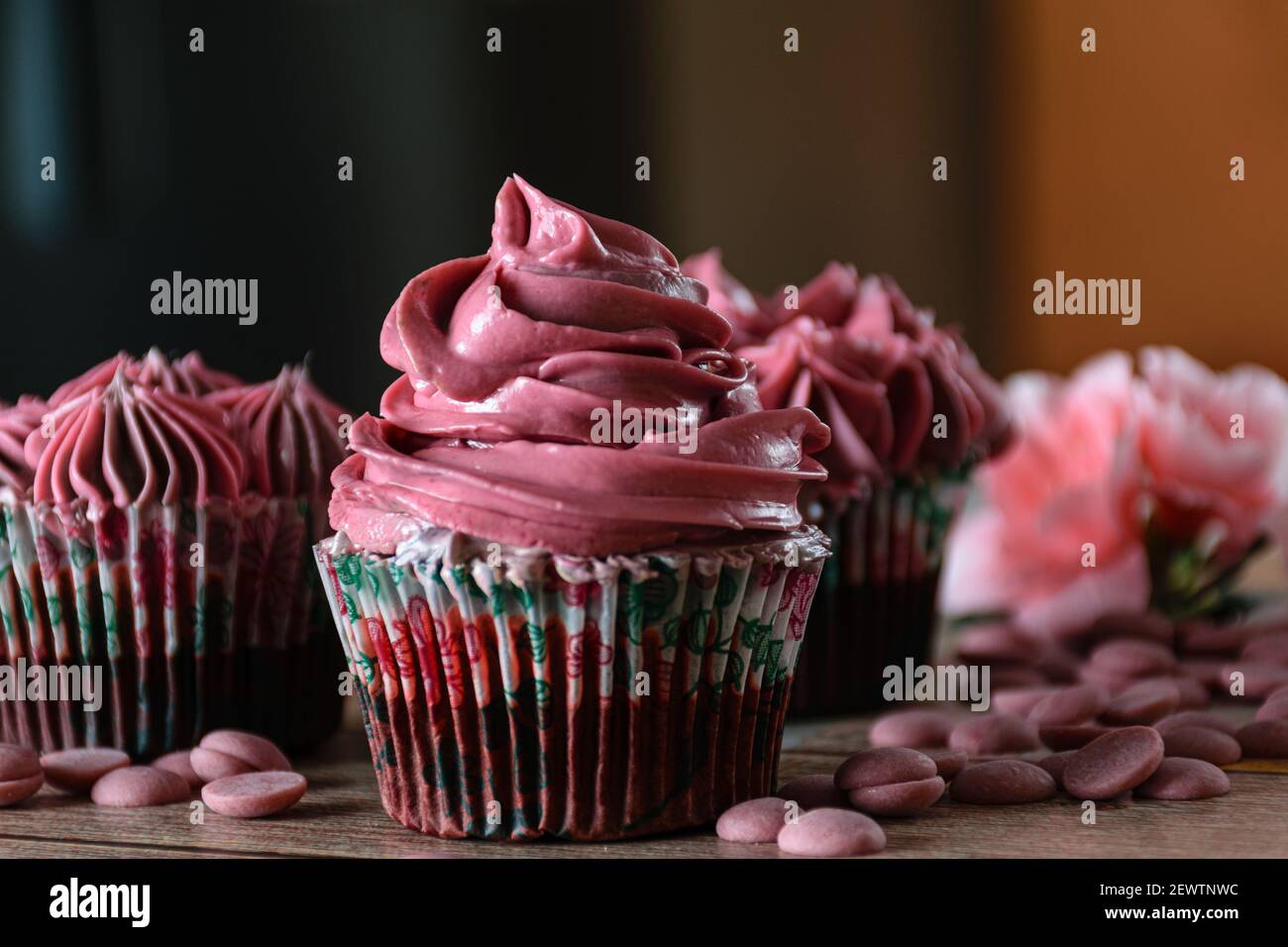 Closeup of three chocolate cupcakes with pink butter cream. Next to Ruby chocolate callets. Brown and orange background. Stock Photo