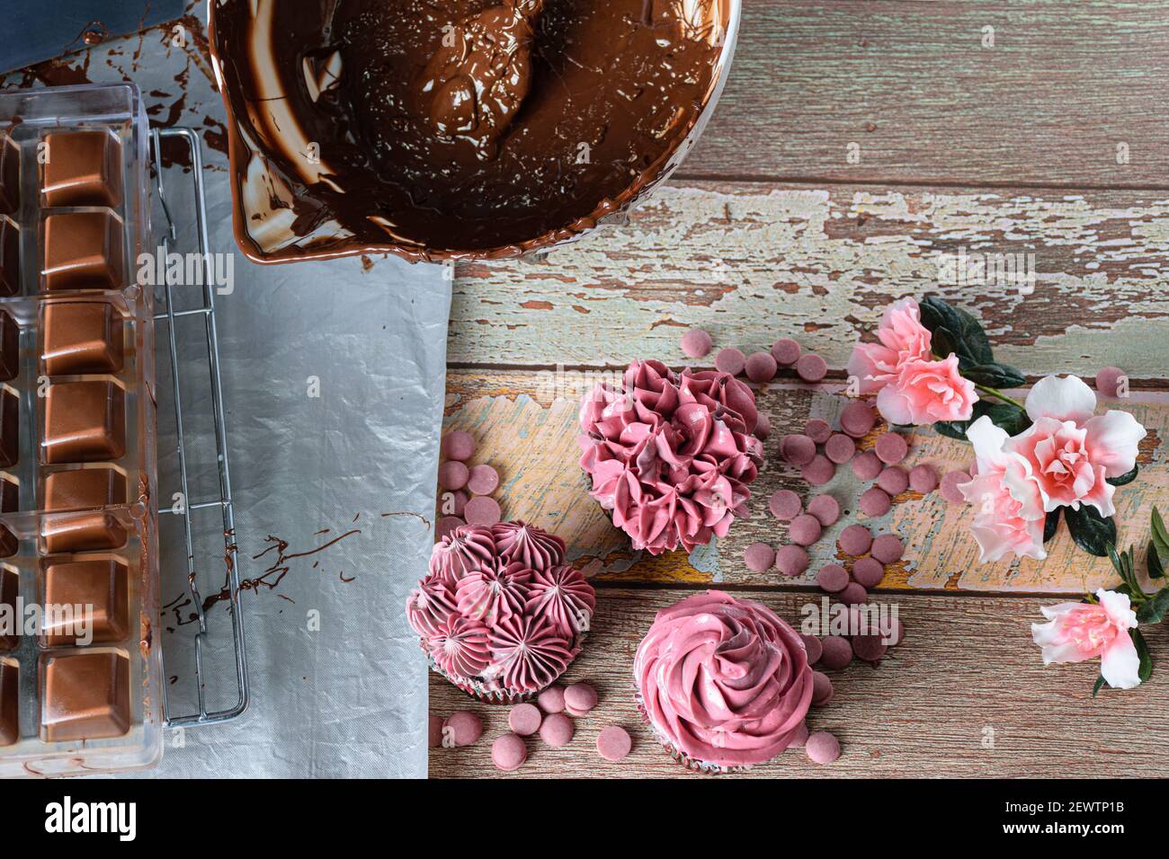 Chocolate cupcakes with pink butter cream, next to ruby chocolate collets, tempered chocolate and a form of pilicarbonate. Stock Photo