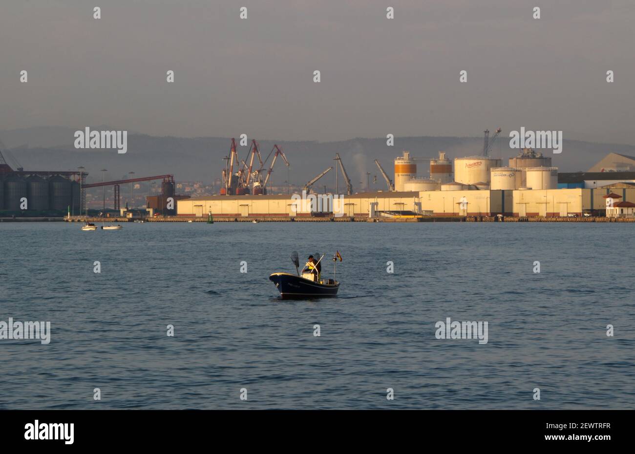 https://c8.alamy.com/comp/2EWTRFR/small-boat-with-lone-person-heading-out-to-fish-in-santander-bay-cantabria-spain-early-morning-with-dock-cranes-silos-and-wharves-behind-2EWTRFR.jpg