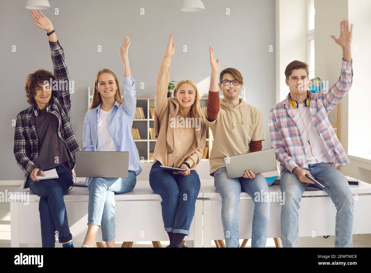 Group of happy active school or college students sitting on desk and raising hands Stock Photo