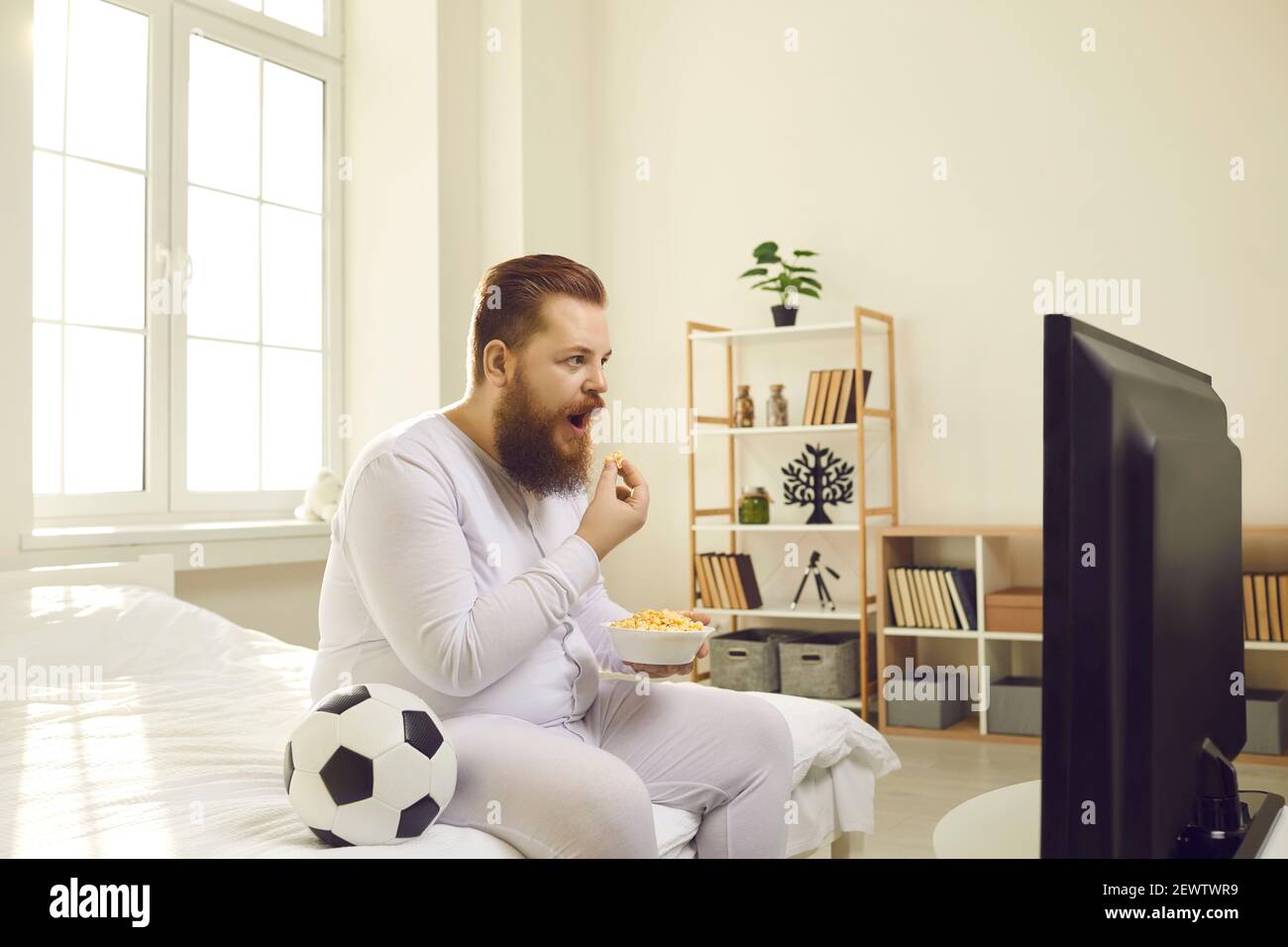 Funny lazy fat sports fan in pyjamas sitting on bed and watching