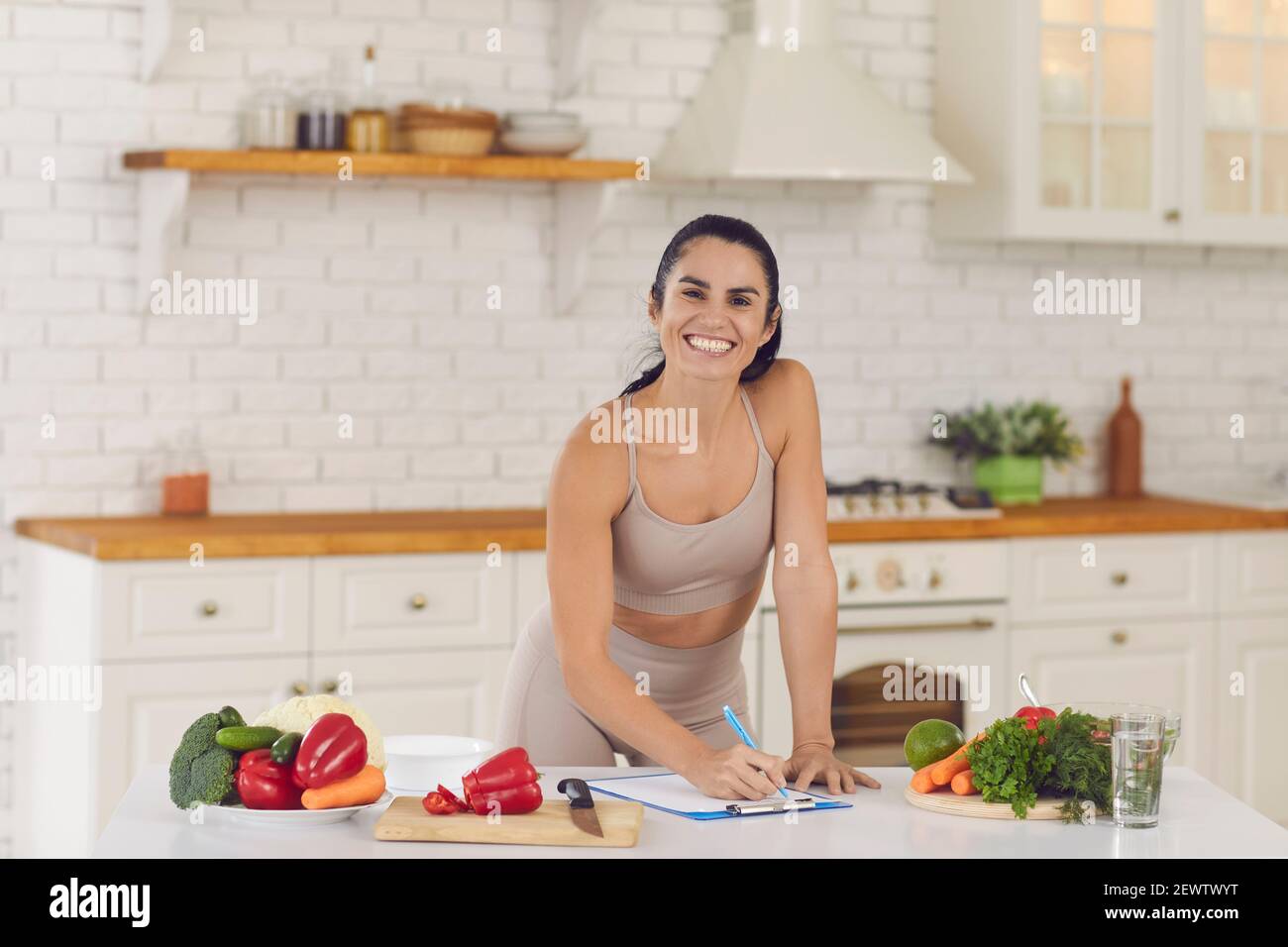 Smiling fitness woman in sportswear standing in kitchen and writing down healthy recipe Stock Photo