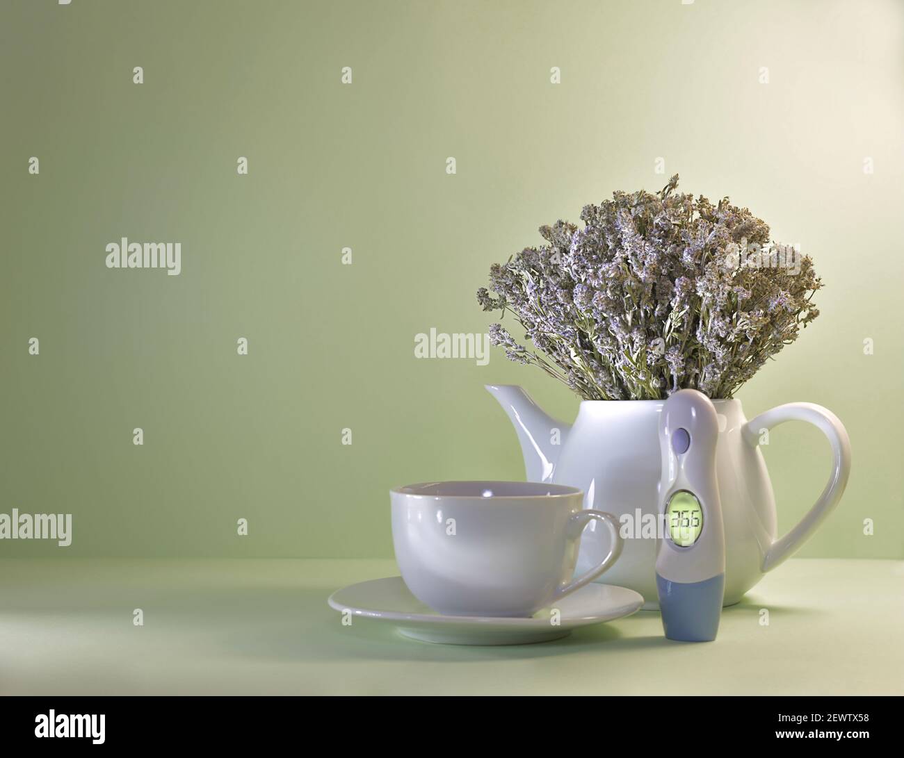 A teapot with a bouquet of dried thyme, next to it is a cup and saucer and a thermometer with a luminous screen. On a light green background. Stock Photo