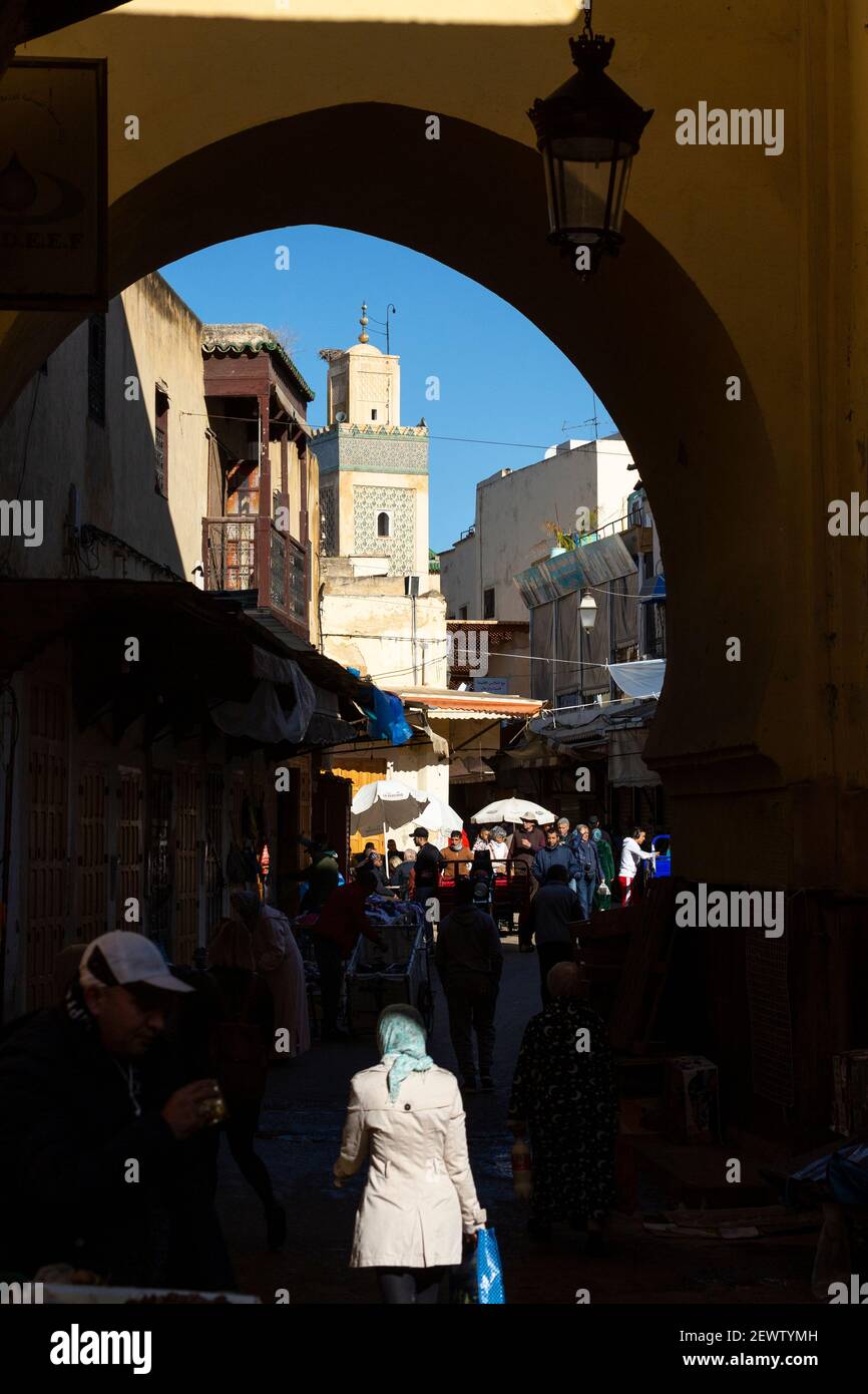 A woman walks into shadow beneath a medieval gate with minaret in background, Fes, Morocco Stock Photo