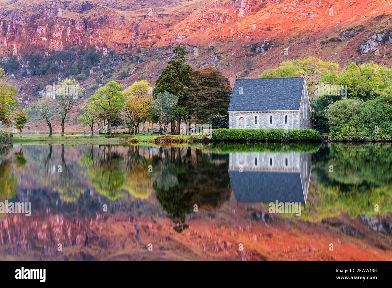 Sunrise lights up the mountains in autumn colours behind St Finbarr's Oratory and reflects in the lake at Gougane Barra, West Cork, Ireland. Stock Photo
