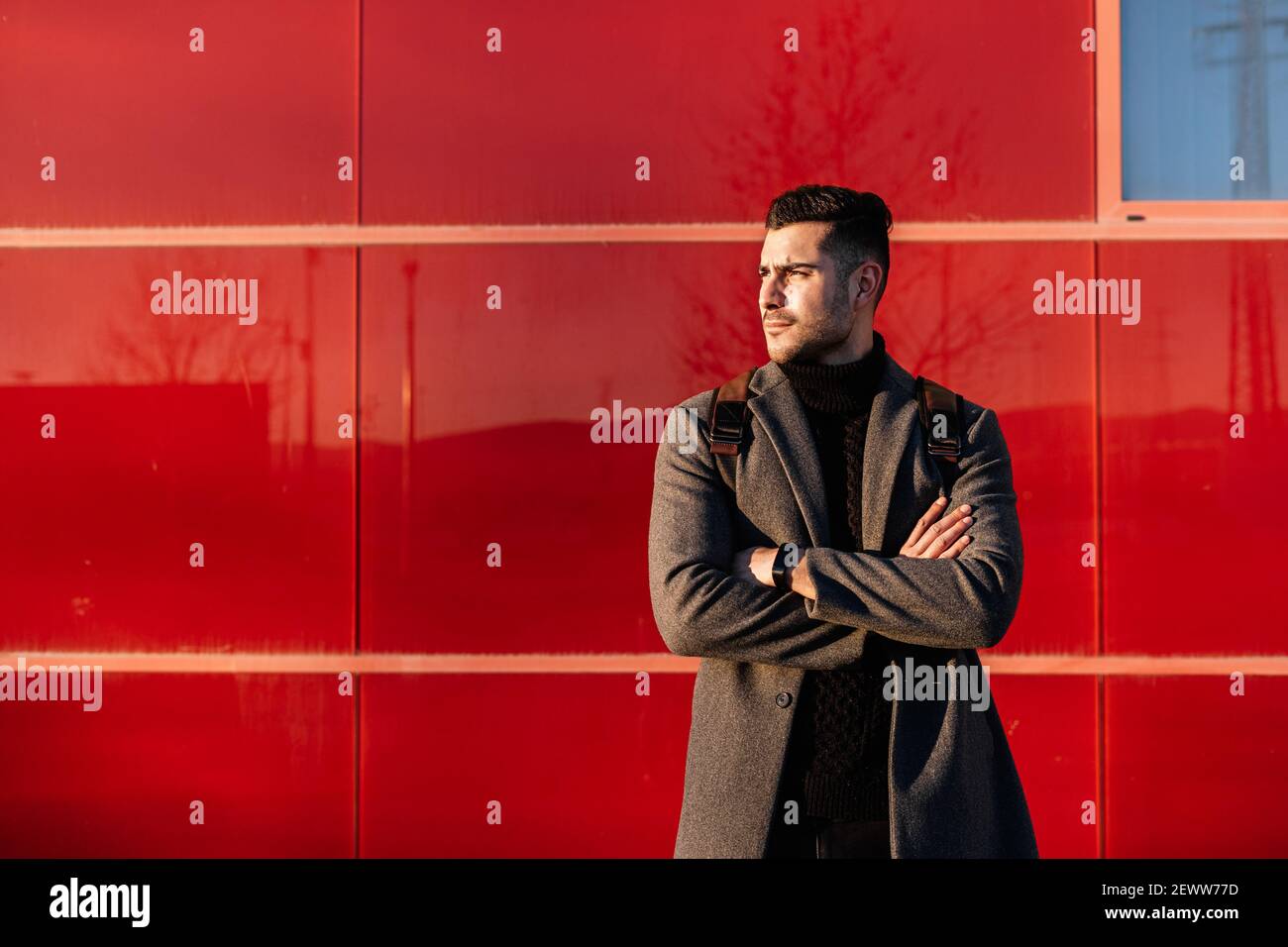 Young businessman with backpack and arms crossed in front of a red wall at sunset. Stock Photo