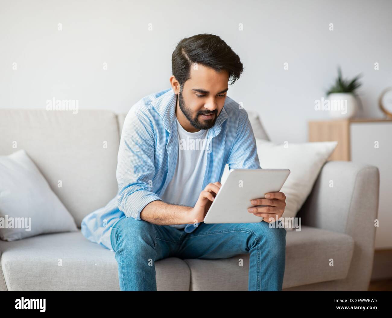 Handsome Eastern Guy Using Digital Tablet While Relaxing On Couch At Home Stock Photo