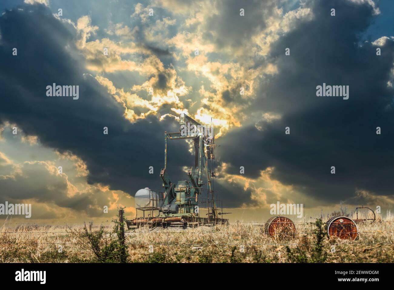Oil well pump jack in field with old oil barrels behind barbed wire fence under dramatic sunset sky with dark oninous clouds Stock Photo