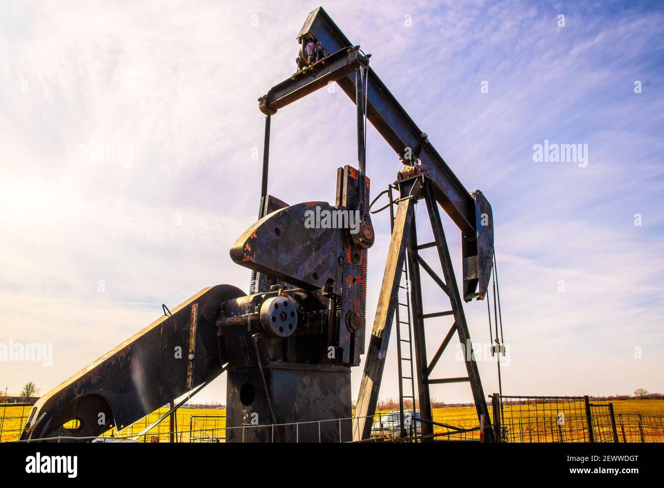 Old rusty oil well pump jack on winter day under strange streaked puple sky in winter pasture with a car parked behind it Stock Photo