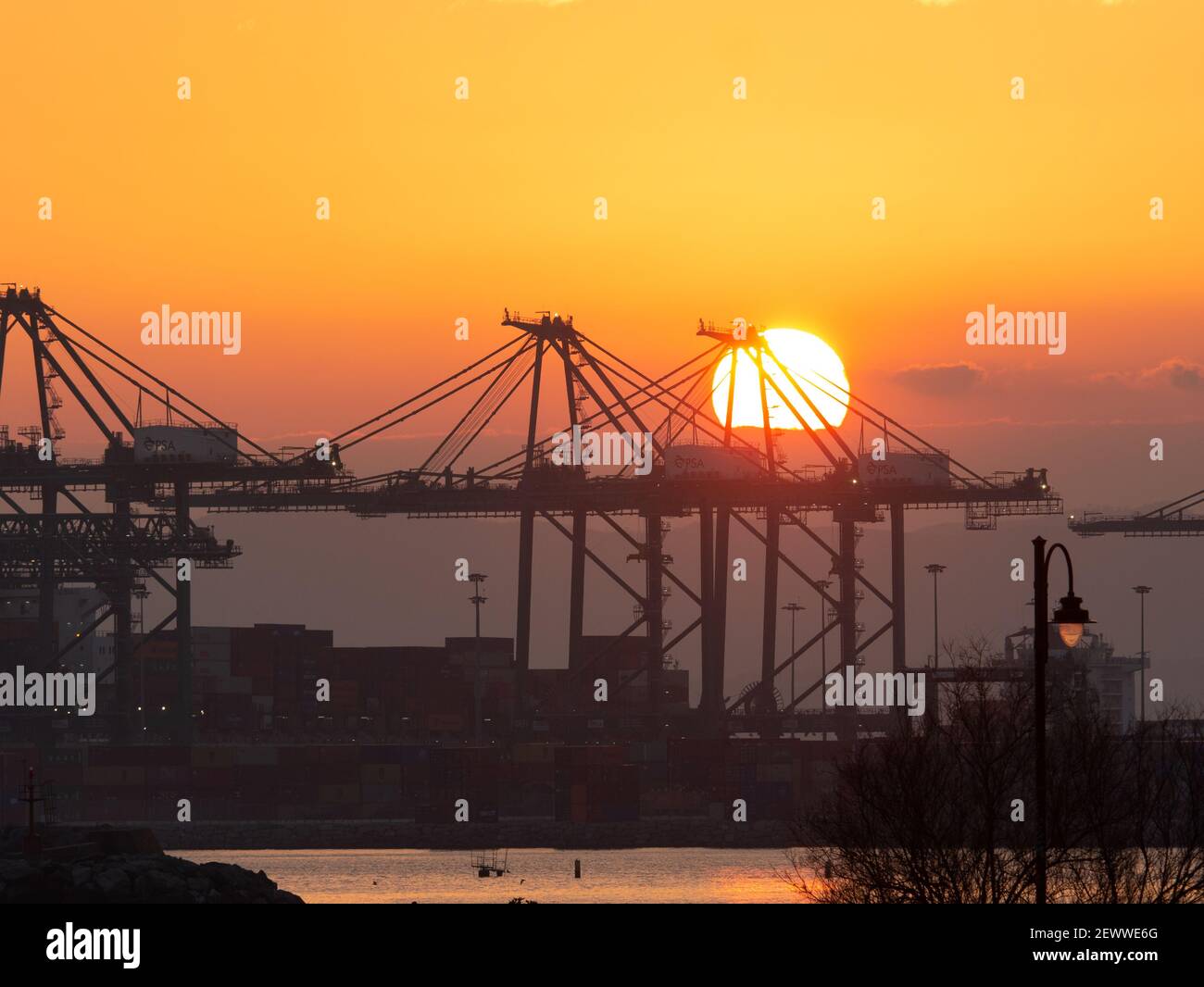 The Sun Sets Between The Large Cranes Of The Port Used To Load And the-sun-sets-between-the-large-cranes-of-the-port-used-to-load-and