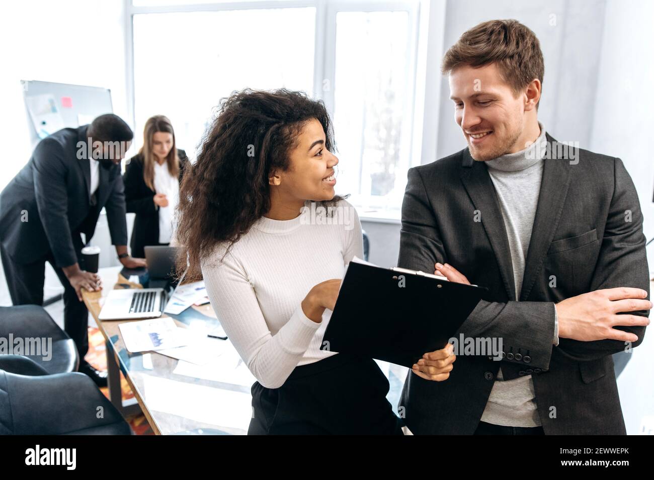 Two multiracial successful colleagues communicate in a modern office, discuss new ideas, think about a plan, in the background their colleagues Stock Photo