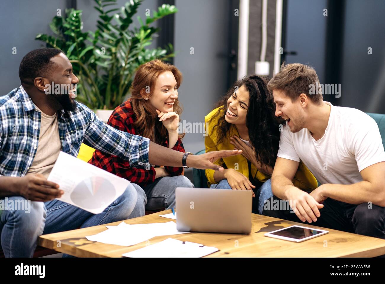 Joyful friendly employees or students take a break from work or study, sitting on the sofa, telling funny stories. Multiracial male and employee coworkers are having a pleasant conversation, laughing Stock Photo