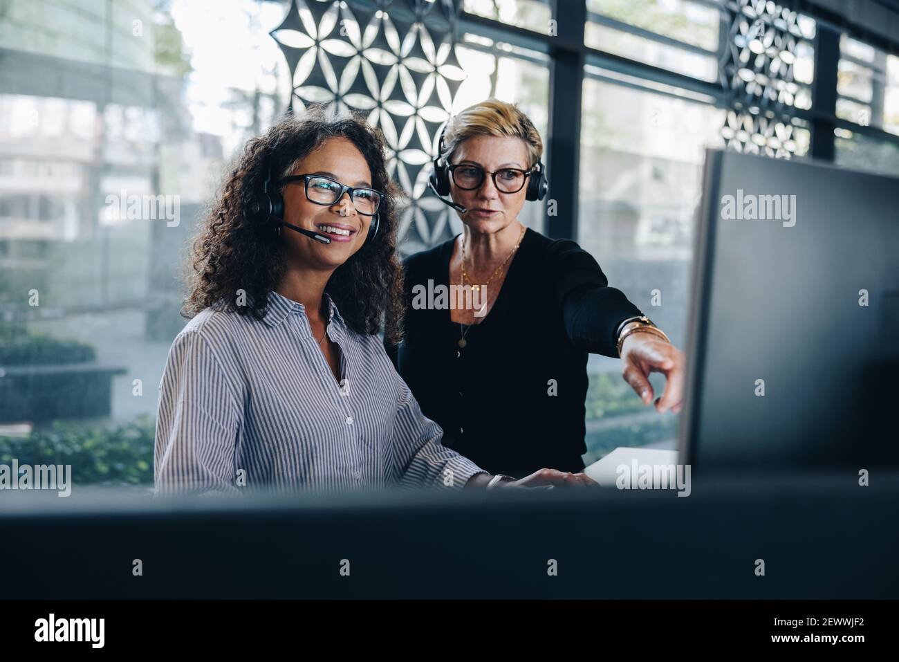 Two businesswomen cooperating while using computer and working in the office. Manager helping female colleague working on computer. Both wearing heads Stock Photo