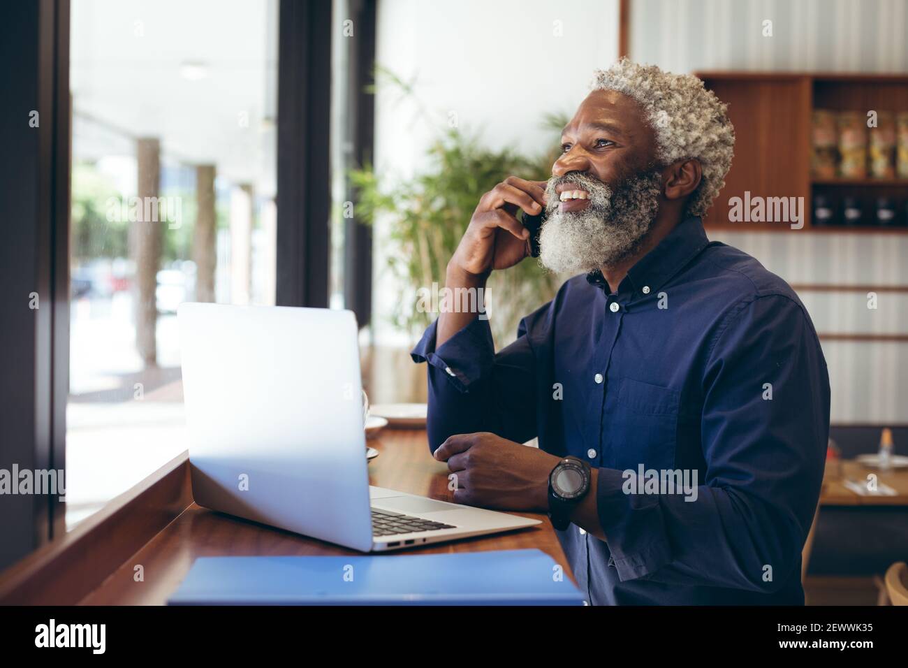 African american senior man sitting at table in cafe using laptop talking on smartphone and smiling Stock Photo