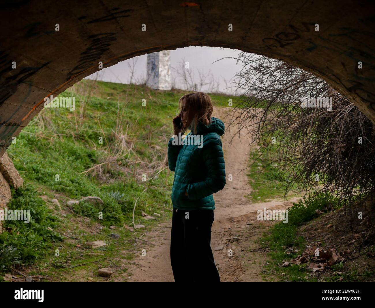 A blonde female in a green winter coat talking on the phone in a tunnel under a road crossing Stock Photo