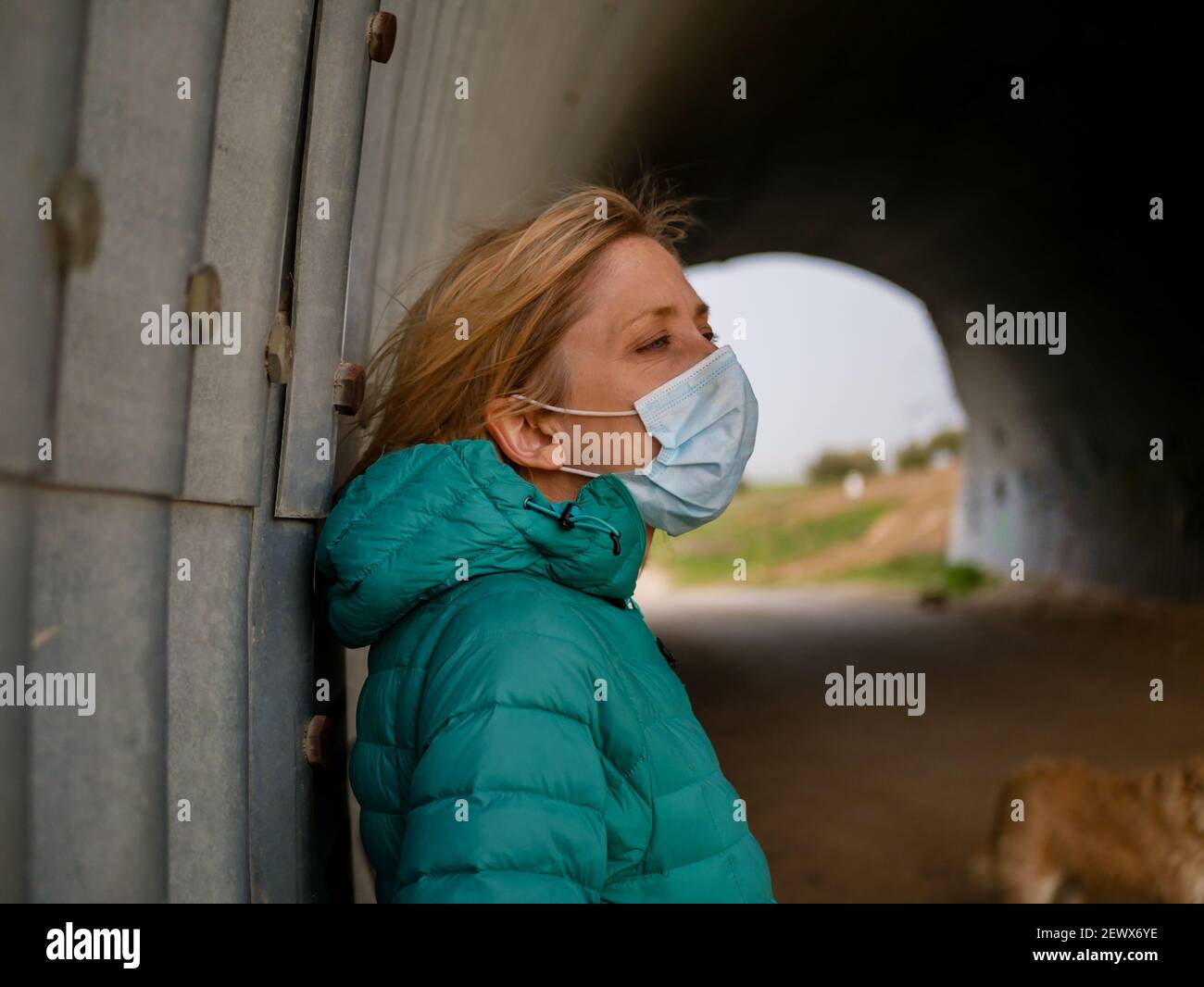 A blonde female wearing a mask posing in a tunnel under a road crossing Stock Photo