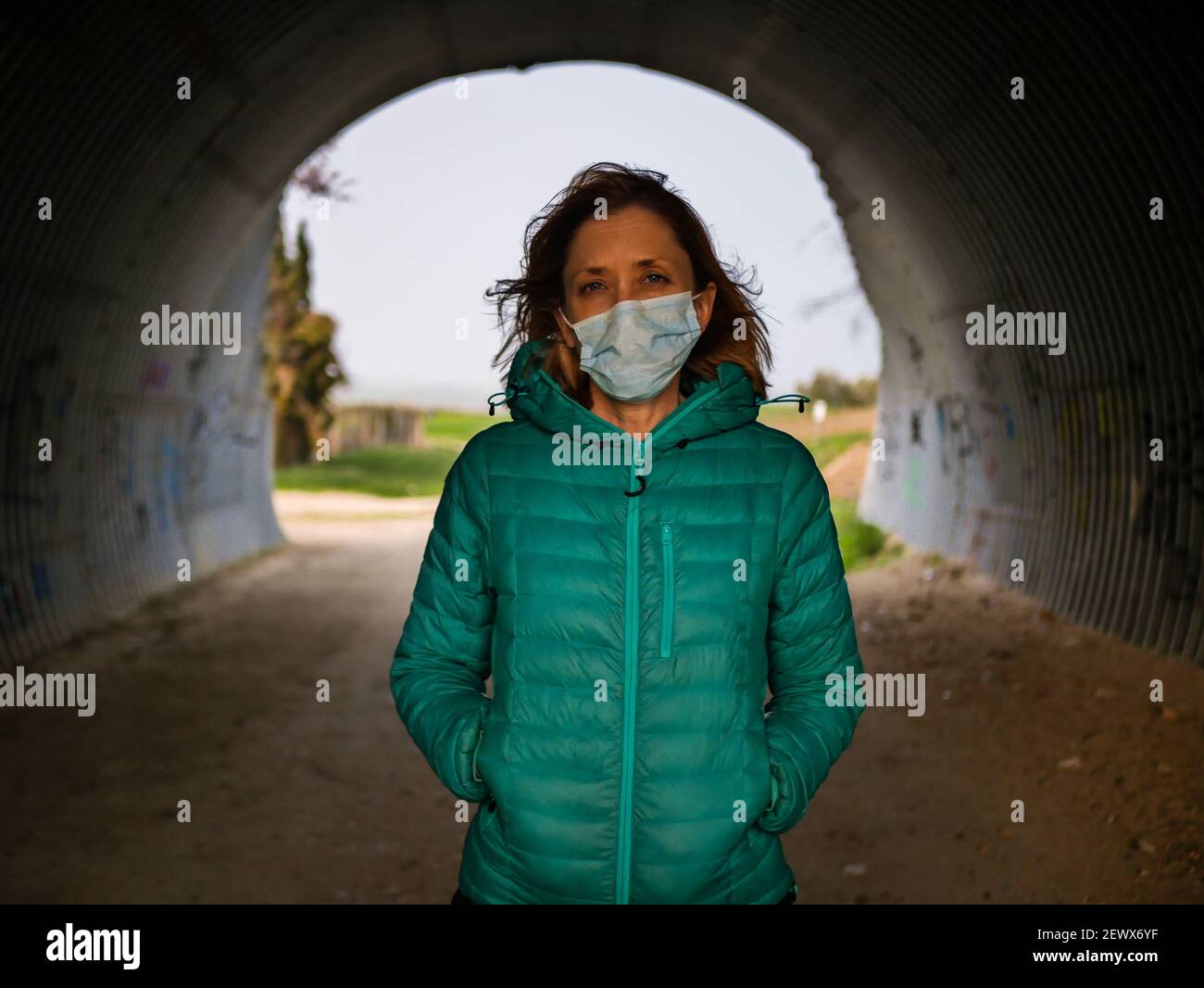 A blonde female wearing a mask posing in a tunnel under a road crossing Stock Photo