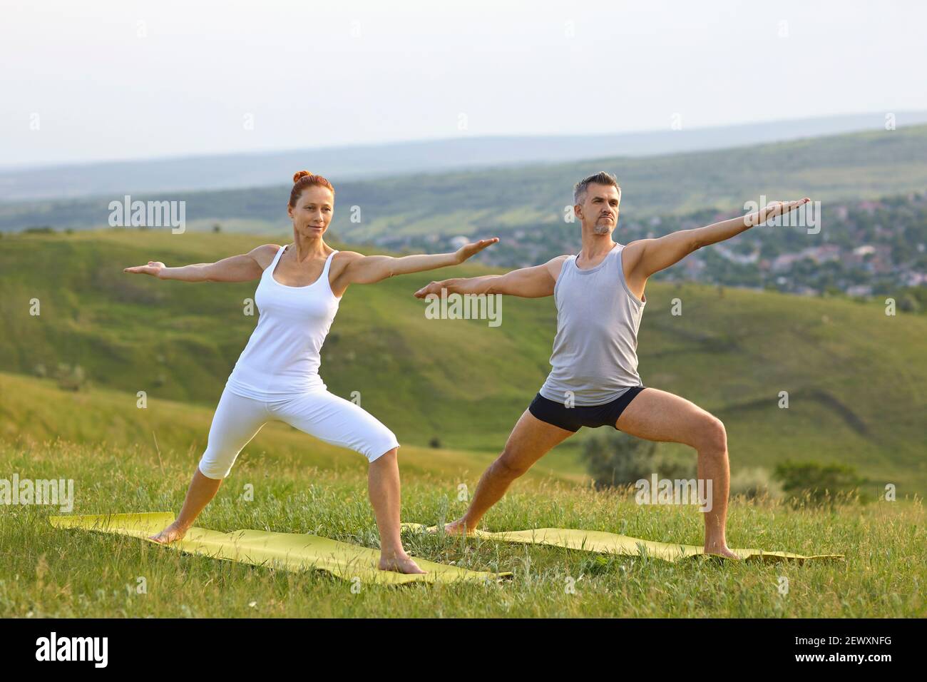 Middle aged couple doing Warrior II pose while practicing yoga on grassy hill in countryside Stock Photo