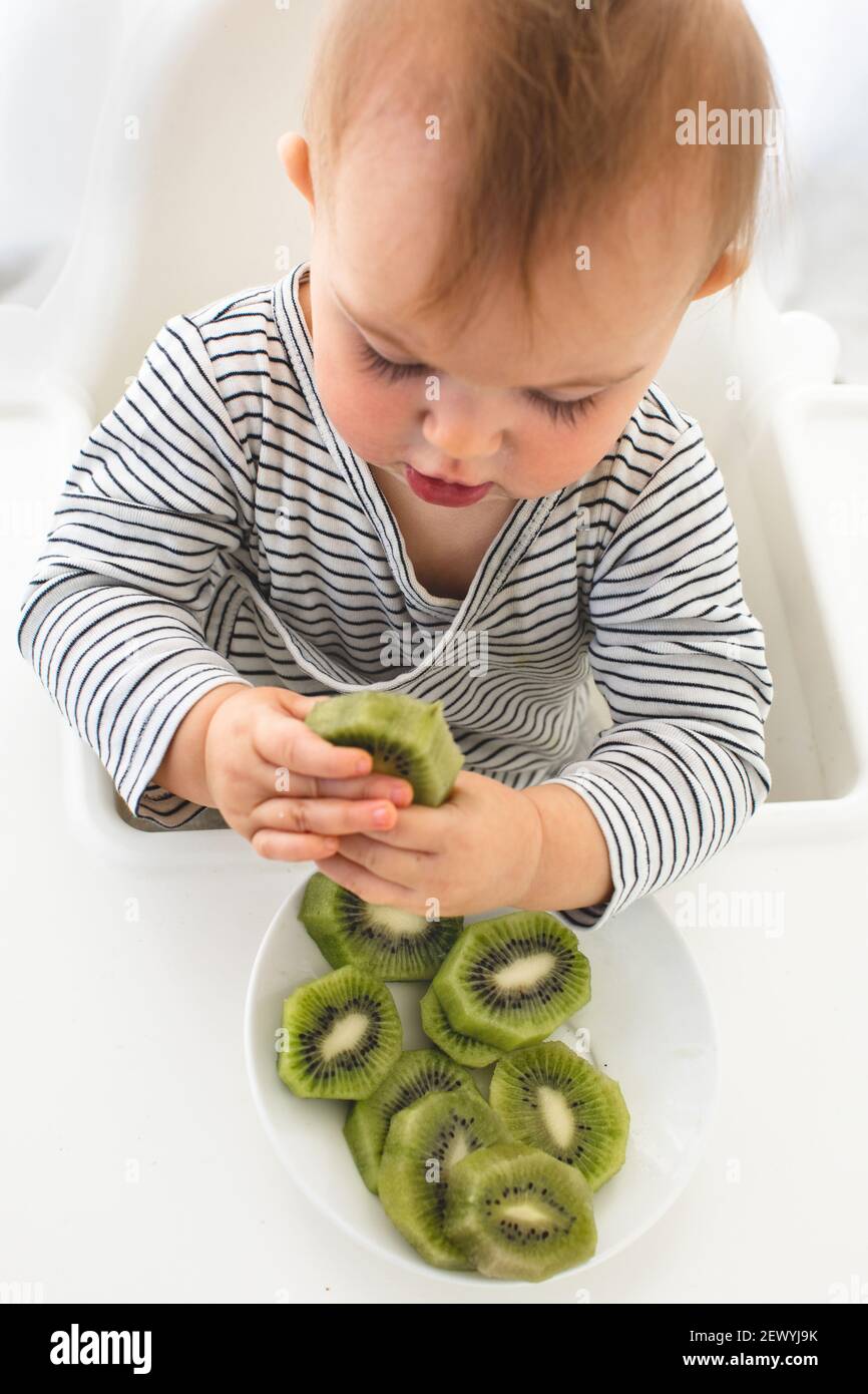 Cute baby girl are sitting whith kiwi white background interior. Funny child explores the fruit Stock Photo