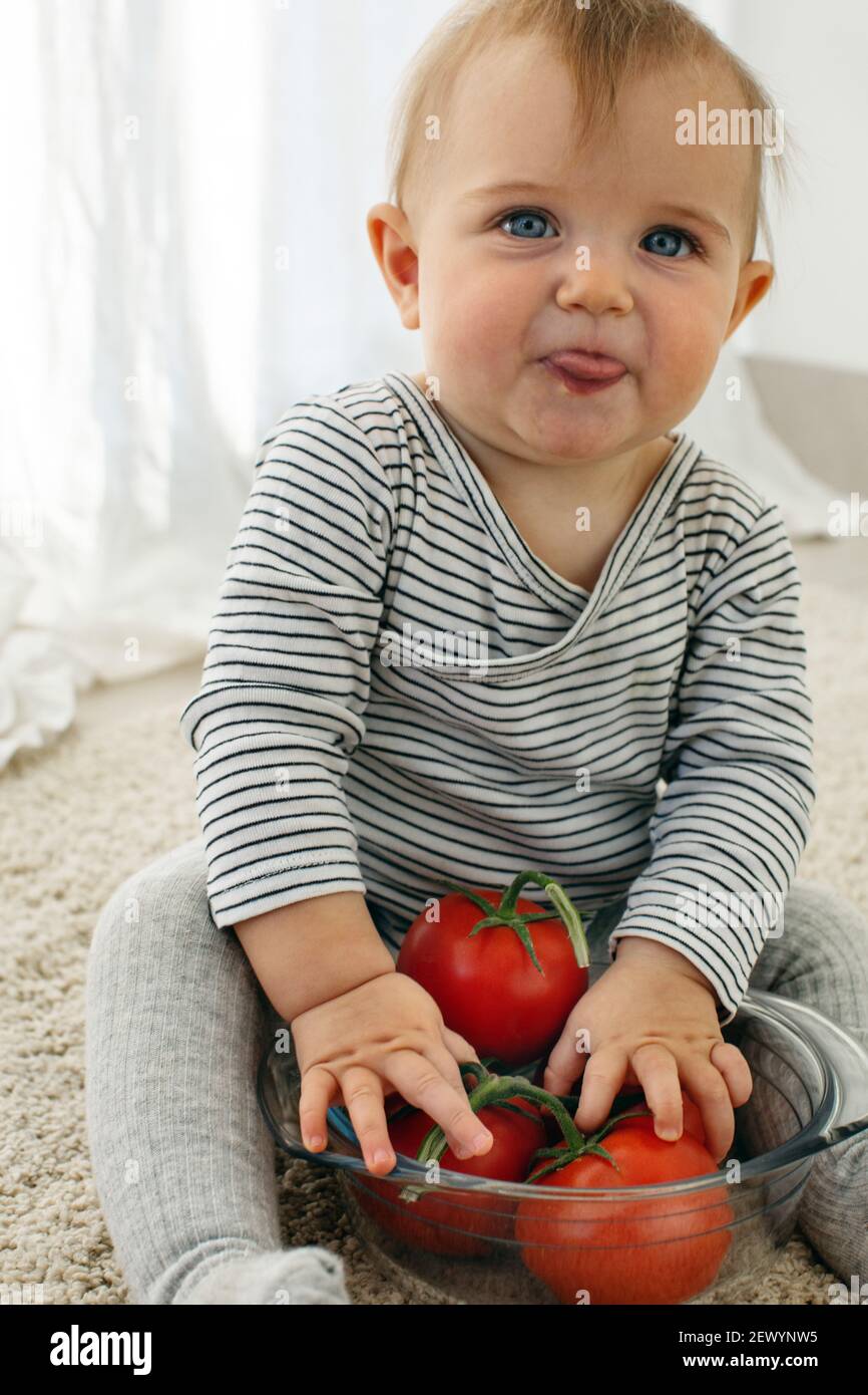 Cute baby girl are sitting whith tomato white background interior. Funny child explores vegetable Stock Photo