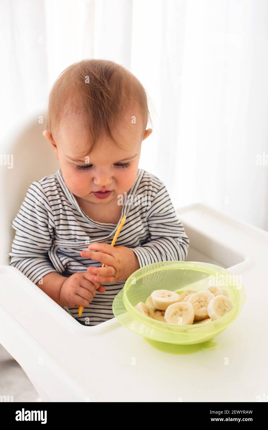 Cute baby girl are sitting whith sliced banana white background interior. Funny child explores the fruit Stock Photo