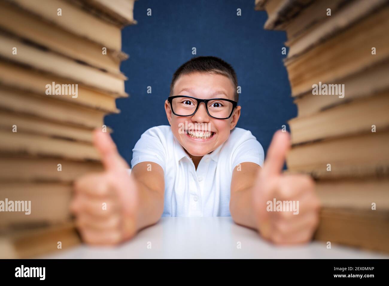 Happy smart boy in glasses sitting between two piles of books and look