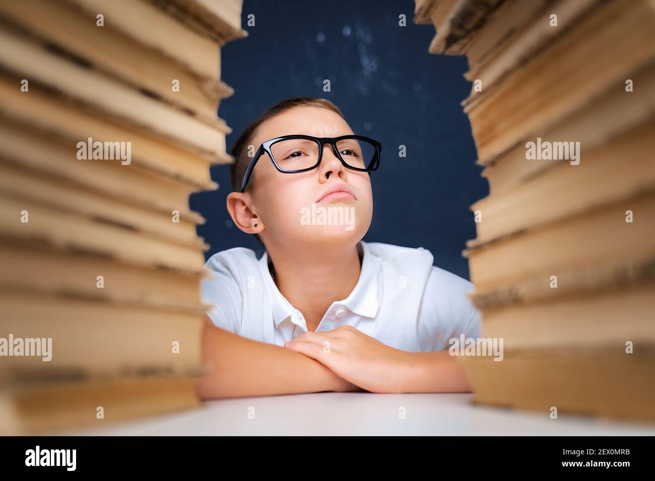 Smart boy in glasses sitting between two piles of books and look up puffed out cheeks. Stock Photo